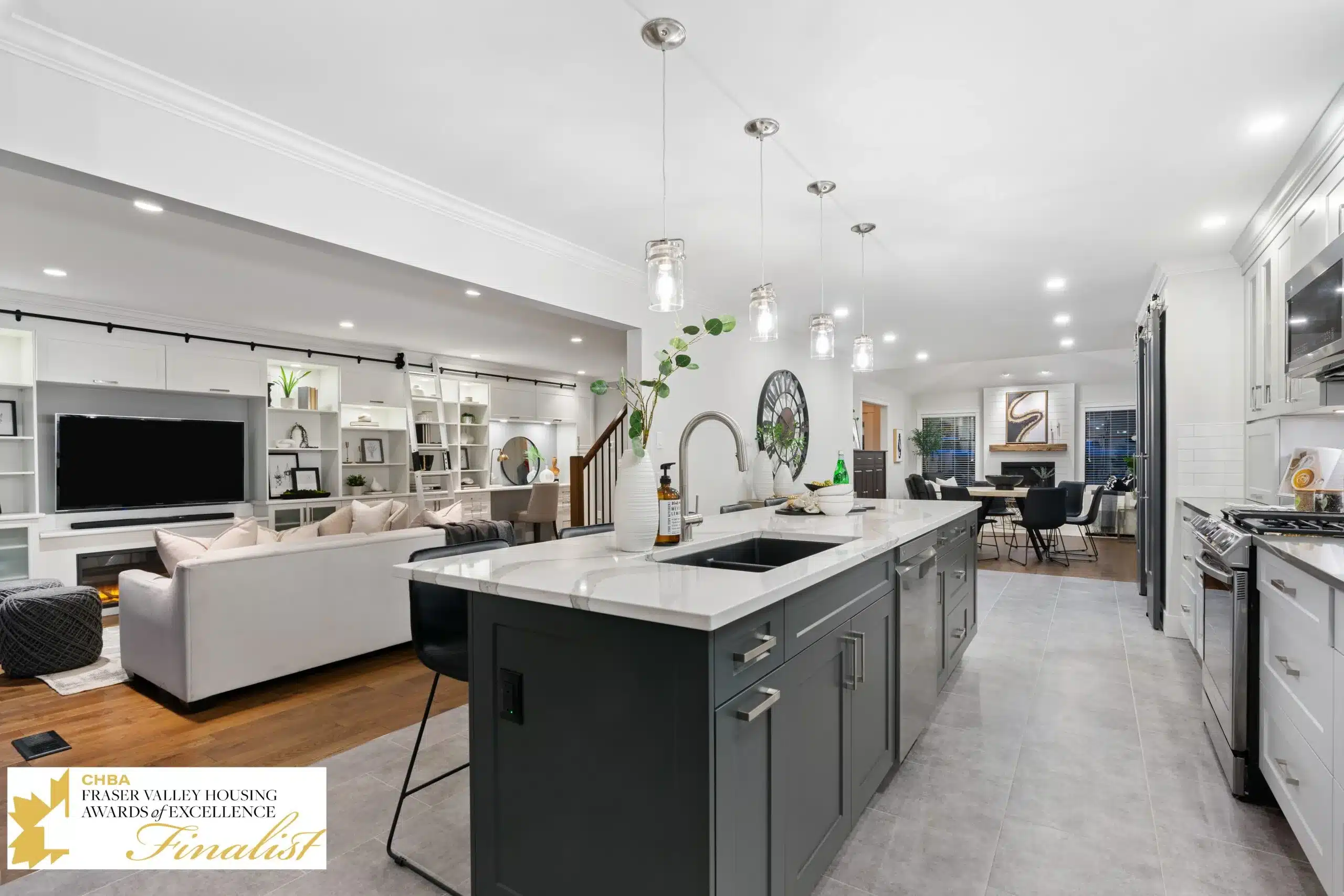 Modern kitchen with gray cabinetry, white countertops, and pendant lighting, featuring an open layout connecting to a stylish living area with a large TV, showcasing multi-functional design and contemporary decor.